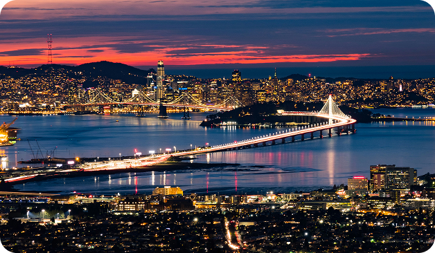 A view of the Bay Bridge illuminated at dusk, connecting San Francisco and Oakland, with city lights and a colorful sunset sky in the background.