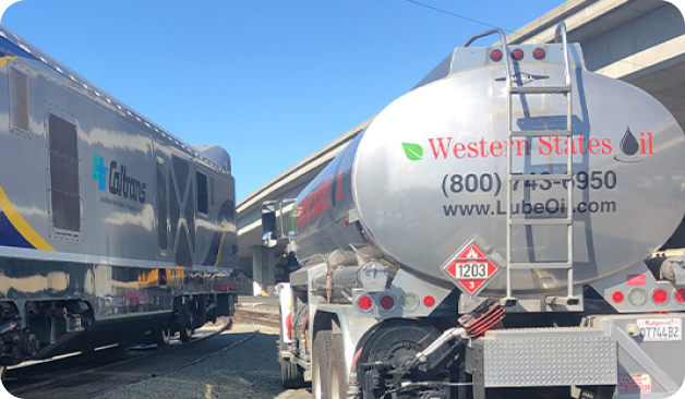 A fuel delivery truck labeled "Western States Oil" is parked near a train marked "Caltrans," with overpasses visible in the background.