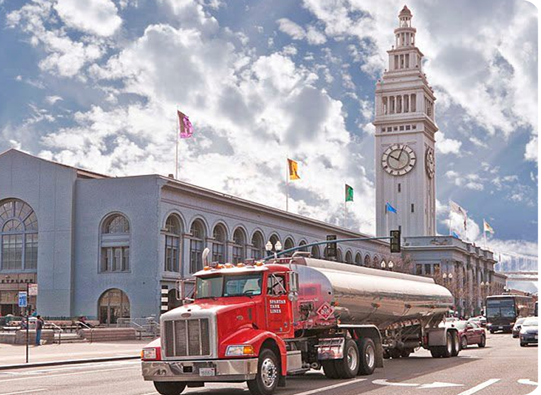 A red fuel tanker truck drives past a large building with a clock tower and flags, under a partly cloudy sky.