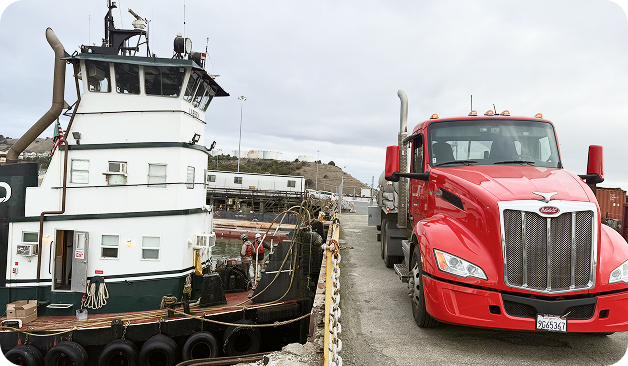 A red semi-truck is parked on a dock next to a moored tugboat, with industrial buildings and equipment visible in the background.
